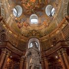 Eine Bibliothek mit hohen Bücherregalen und einer Statue in der Mitte. Die Decke zeigt ein aufwändiges Fresko mit Figuren.