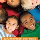 „Zukunft braucht Demokratie. Demokratie braucht Bildung.“ Fünf lachende Kinder liegen im Kreis auf bunten Matten.