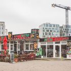 "Arena Bistro" in großen bunten Buchstaben, umgeben von hellen Hochhäusern und einem Kran im Hintergrund.