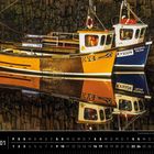 „Schottland 01“ und Text auf Fischerbooten: KY4, KY1004. Zwei bunte Boote spiegeln sich im Wasser im Hafen.