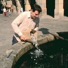 Ein Mann gießt Münzen aus einer Papiertüte in ein steinernes Becken auf einem sonnigen Platz mit wenigen Passanten.