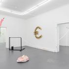 Ein Raum mit weißem Boden und Wänden, hängende bunte Schlauchskulpturen und ein aus Stroh gebildetes Euro-Symbol an der Wand.