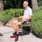 Eine Person sitzt draußen in einem Garten, umgeben von grünen Pflanzen. Sie trägt ein weißes Hemd und rote Schuhe.