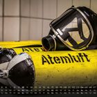 Auf gelbem Zylinder steht „Atemluft 300“. Darauf liegt eine schwarze Atemschutzmaske. Darunter ein Kalender.