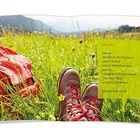 Auf einer grünen Wiese liegen rote Wanderschuhe und ein roter Rucksack. Im Hintergrund sind Berge zu sehen.