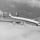 "Lufthansa" auf einem klassischen Propellerflugzeug in der Luft, umgeben von Wolken, mit Firmenlogos auf den Leitwerken.