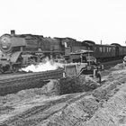 Eine Dampflok fährt schnell auf Schienen vorbei, während ein Bagger Erde neben den Gleisen bewegt.