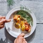 Ein Teller mit geräuchertem Lachs, Dill, Brot und pochiertem Ei. Zwei Hände benutzen Messer und Gabel.