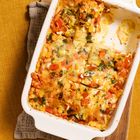 Ein Auflauf in einer weißen Ofenform, serviert auf einem hölzernen Schneidebrett. Enthält rote und grüne Gemüse.