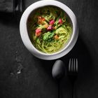 Keine Texte sichtbar. Helles Schälchen mit grüner Pasta, dekoriert mit bunten Toppings; daneben schwarzes Besteck.