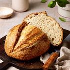 Ein Laib frisch gebackenes Brot auf einem Holzbrett, daneben ein Messer mit Holzgriff und ein Kräutertopf.