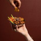 Zwei Hände vor rotem Hintergrund, halten eine Schale mit Streifen von Gemüse und frittierten Stücken; eine Hand greift danach.