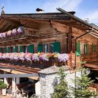 Ein traditionelles Holzhaus in Alpennähe, Balkone mit dicht bepflanzten Blumenkästen, grüne Fensterläden, blauer Himmel.