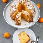 Ein Gugelhupf mit Mandeln, bestäubt mit Puderzucker. Ein Stück ist geschnitten auf einem Teller. Aprikosen liegen daneben.