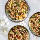 Drei Teller mit cremigem Dip, verziert mit Pinienkernen, Frühlingszwiebeln und Soße. Daneben zwei Gläser mit hellgelbem Getränk.