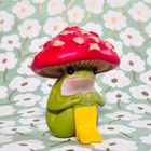 Ein grüner Frosch mit gelben Stiefeln und einem roten Pilzhut sitzt vor einem Blumenhintergrund.