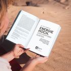 Text: "Energie hoch. Erschöpfung runter." Eine Person liest ein Buch am Strand, umgeben von Sand.