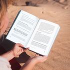 Ein aufgeschlagenes Buch wird am Strand gehalten, die Seiten sind mit Text gefüllt. Eine Person hält es, Blick auf Sand.