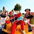 Text „Flötzi“ auf Cheerleader-Outfits. Mann in rotem Anzug und Kappe posiert vor gelbem Auto, begleitet von drei Frauen.