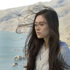 Es gibt keinen Text zu beschreiben. Eine Frau steht im Wind mit langen Haaren und schaut ernst; im Hintergrund Berge und Meer.