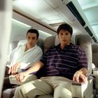 Zwei Männer sitzen in einem Flugzeug. Links trägt einen weißen Hemd, rechts dunkles Polohemd. Eine Flugbegleiterin ist rechts hinten.