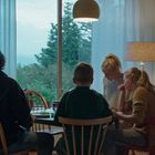 Vier Personen sitzen an einem Tisch in einem Wohnzimmer, das von einer großen Fensterfront erleuchtet wird.