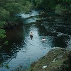 Zwei Menschen schwimmen in einem dunklen Fluss, umgeben von dichtem Grün.