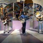 Eine Band spielt auf einer bunten Bühne mit psychedelischen Mustern. Musiker tragen bunte Kleidung.