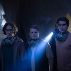 Drei junge Männer mit Atemmasken stehen in einem düsteren Raum, erleuchtet von einer Taschenlampe.