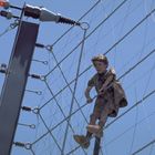 Ein Kind klettert an einem hohen, elektrifizierten Zaun mit Drähten, der Himmel ist klar und blau.