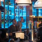 Zapfhähne mit Markenlogos vor blau beleuchtetem Regal; Menschen lachen an einer Bar.