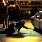 Ein Mann kriecht auf einem Bürgersteig auf ein parkendes Polizeiauto zu, während umstehende Personen zusehen.