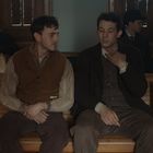 Zwei Männer sitzen auf einer Holzbank in einem wartesaalähnlichen Raum. Einer hält seine Hand am Hals.