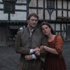 Ein Paar in mittelalterlicher Kleidung steht lächelnd vor einem Fachwerkhaus. Im Hintergrund sind Menschen und ein Hund.