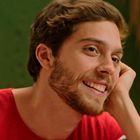 Ein Mann mit braunen Haaren und Bart lächelt. Er trägt ein rotes Shirt. Der Hintergrund ist grünlich.