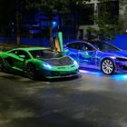 Ein grüner Sportwagen und ein blau-leuchtender Wagen parken nachts auf einer Straße, umgeben von Bäumen.