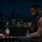 Eine Frau und ein Mann sitzen an einer Bar. Sie reden, während er ein Glas hält. Im Hintergrund eine beleuchtete Skyline.