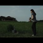 Keine erkennbaren Texte. Zwei Personen auf einer Wiese, eine hält eine Urne. Im Hintergrund ein Haus.