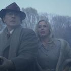 Ein Mann in Mantel und Hut mit einer Frau in Pelzjacke im Freien. Beide schauen besorgt, es ist neblig und bewaldet.