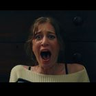 Eine Frau mit erschrockenem Ausdruck, offener Mund, vor einem dunklen Möbelstück. Ihr Pullover ist weiß.