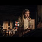Eine Frau mit erschrockenem Blick hält ein aufgeschlagenes Buch mit Abbildungen und Text vor einem liegenden Mann.