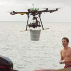„MERCURY“ auf einem Außenbordmotor. Ein Mann ohne Hemd steuert eine Drohne mit einem Eimer darüber vor einer Meereskulisse.