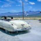 Ein altes Cabrio fährt auf einer staubigen Straße. Spielkarten fliegen heraus. Zwei Personen sitzen darin. Berge am Horizont.