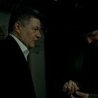 Zwei Männer in einem dunklen Flur, einer im Anzug, der andere mit Mütze und Jacke, tauschen Gegenstände aus.