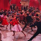 Tanzende Menschen in einem Saal mit roter Wand, in dynamischen Posen, bunt gekleidet, lebendige und kraftvolle Atmosphäre.