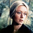 Eine Frau mit blondem Haar schaut nachdenklich zur Seite, mit einem Wald im Hintergrund.