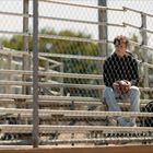 Keine Texte sichtbar. Ein Junge sitzt allein auf leeren Tribünen mit einem Ball in den Händen hinter einem Maschendrahtzaun.