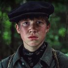 Junger Mann schaut ernst mit rotem Gesicht, trägt Mütze und Schal, Hintergrund ist unscharfer grüner Wald.