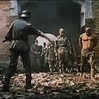 Es sind keine Texte im Bild erkennbar. Soldaten stehen in einer zerstörten Backsteinumgebung und scheinen Häftlinge zu bewachen.