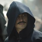 Ein Mann mit Schnurrbart trägt eine dunkle Kapuze und blickt nach rechts. Die Umgebung ist unscharf.